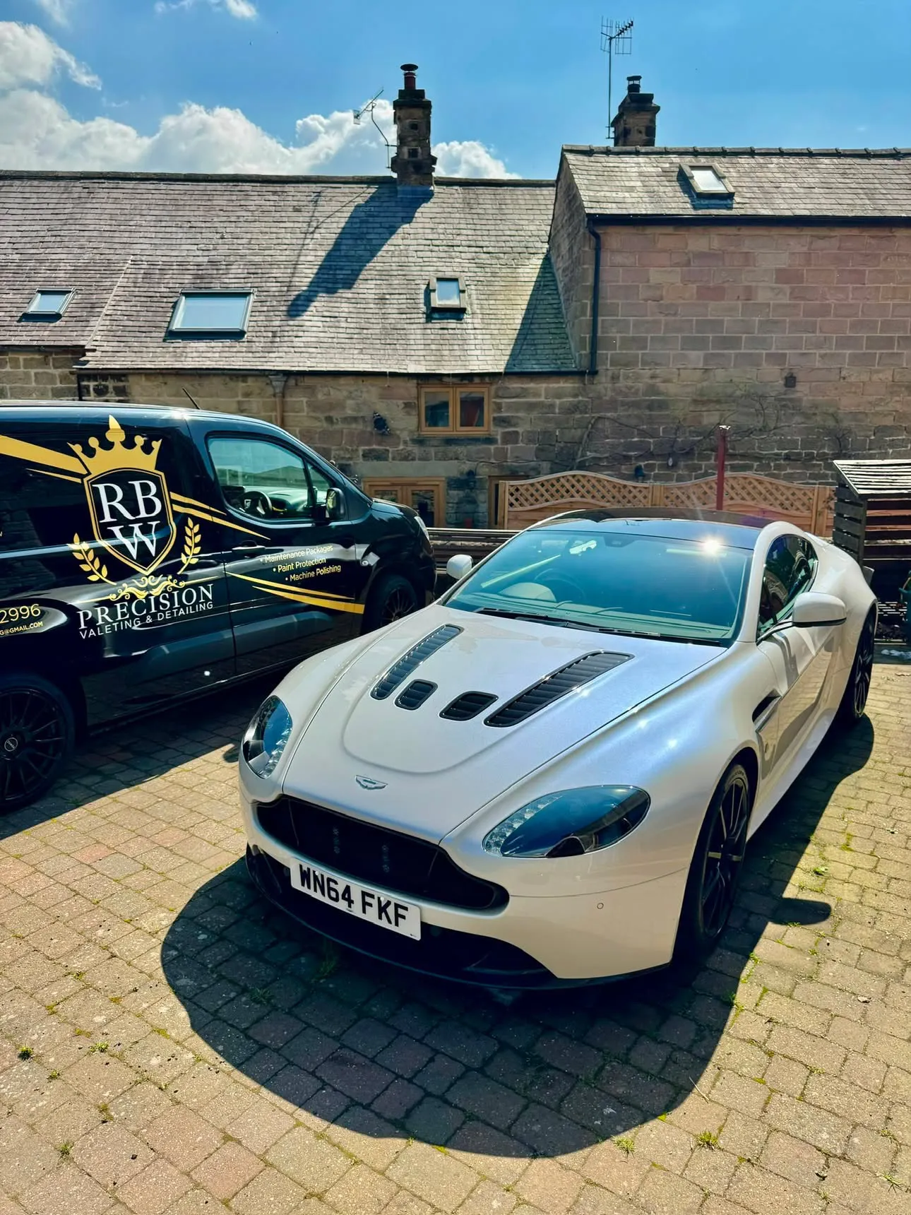 White Aston Martin V12 Vantage on a stone-flagged courtyard, bonnet gleaming in the sun, with the RBW Precision van parked alongside.