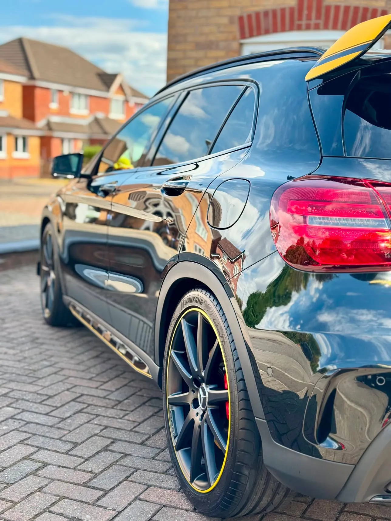 Black Mercedes-AMG GLA45 with yellow accents on a block-paved driveway, the sky and clouds reflected sharply in the freshly finished paintwork.