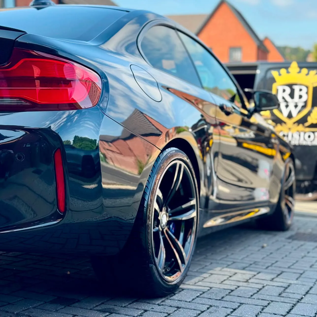 Black BMW M2 close up over the rear wheel, evening sun in the paint, the gold RBW crest on the van behind.